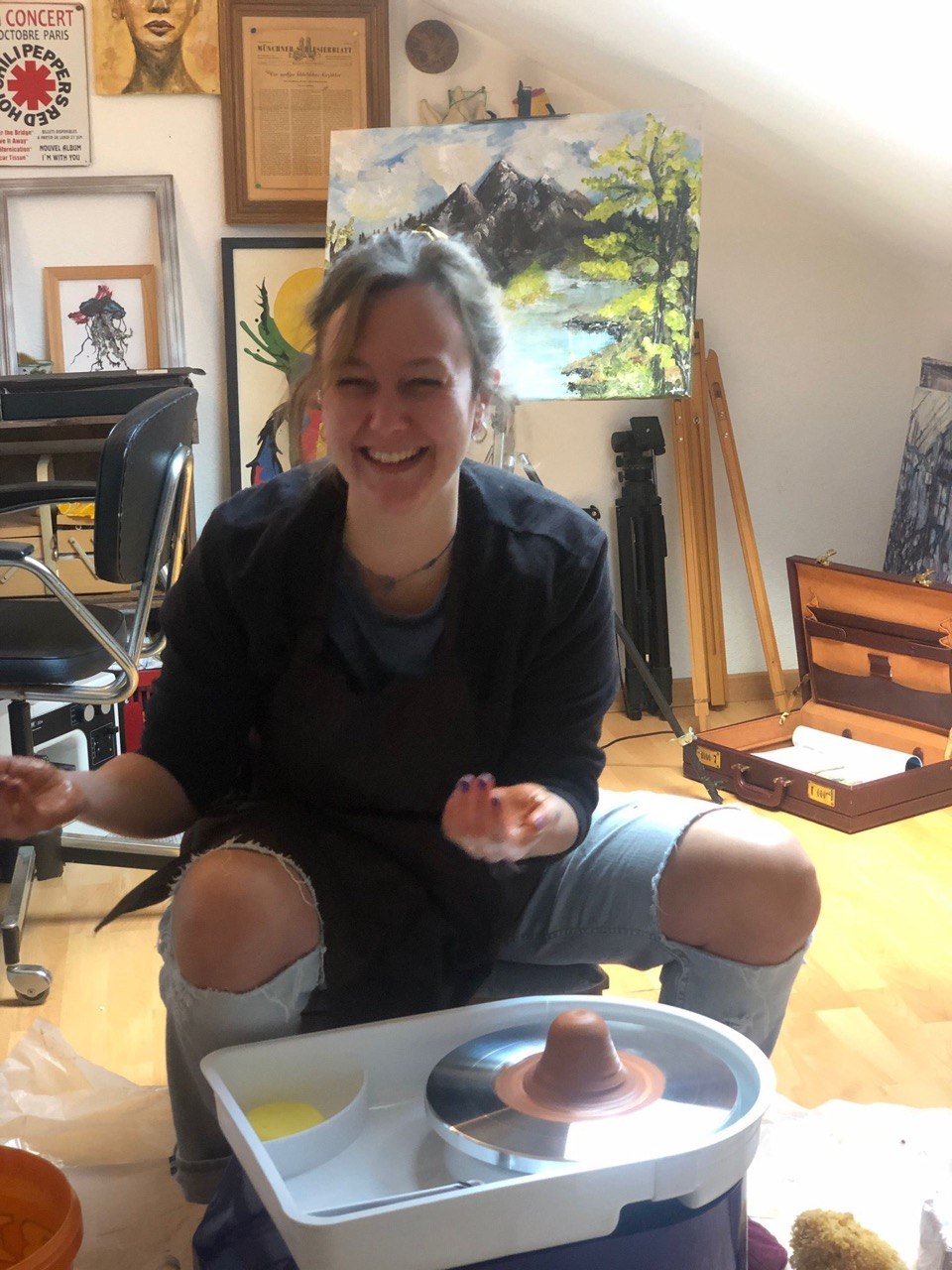 Ein Bild von Lisa in ihrem Atelier wie sie lächelnd zwischen Staffelei und Farbe an ihrer Töpferscheibe.